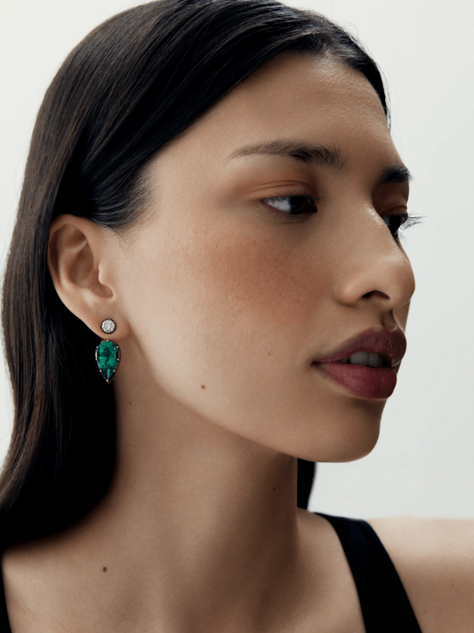 Close-up of a woman wearing elegant stud earrings featuring a pear-shaped emerald and a round diamond, showcasing contemporary design with antique-inspired craftsmanship. View 3