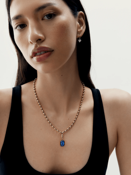 Close-up of a woman wearing a contemporary rose gold necklace featuring a deep blue emerald-cut sapphire pendant, paired with minimalistic silver drop earrings, styled with a black sleeveless top.