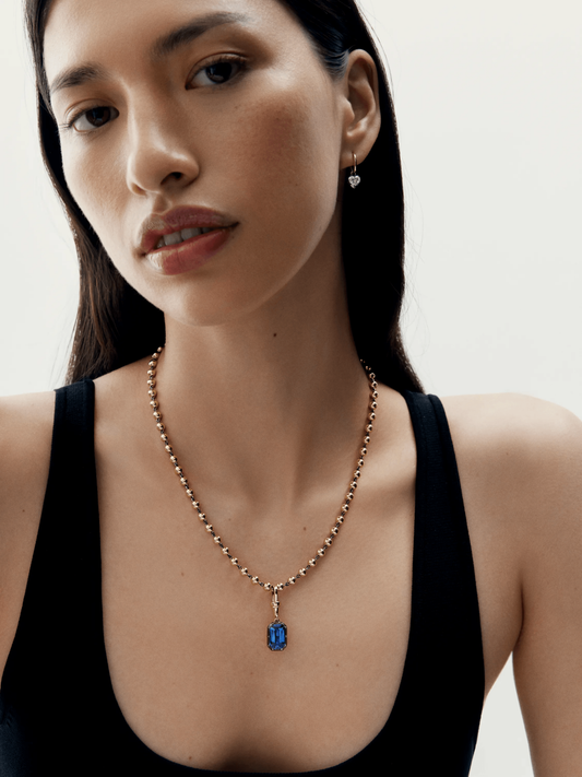 Close-up of a woman wearing a contemporary rose gold necklace featuring a deep blue emerald-cut sapphire pendant, paired with minimalistic silver drop earrings, styled with a black sleeveless top. View 3