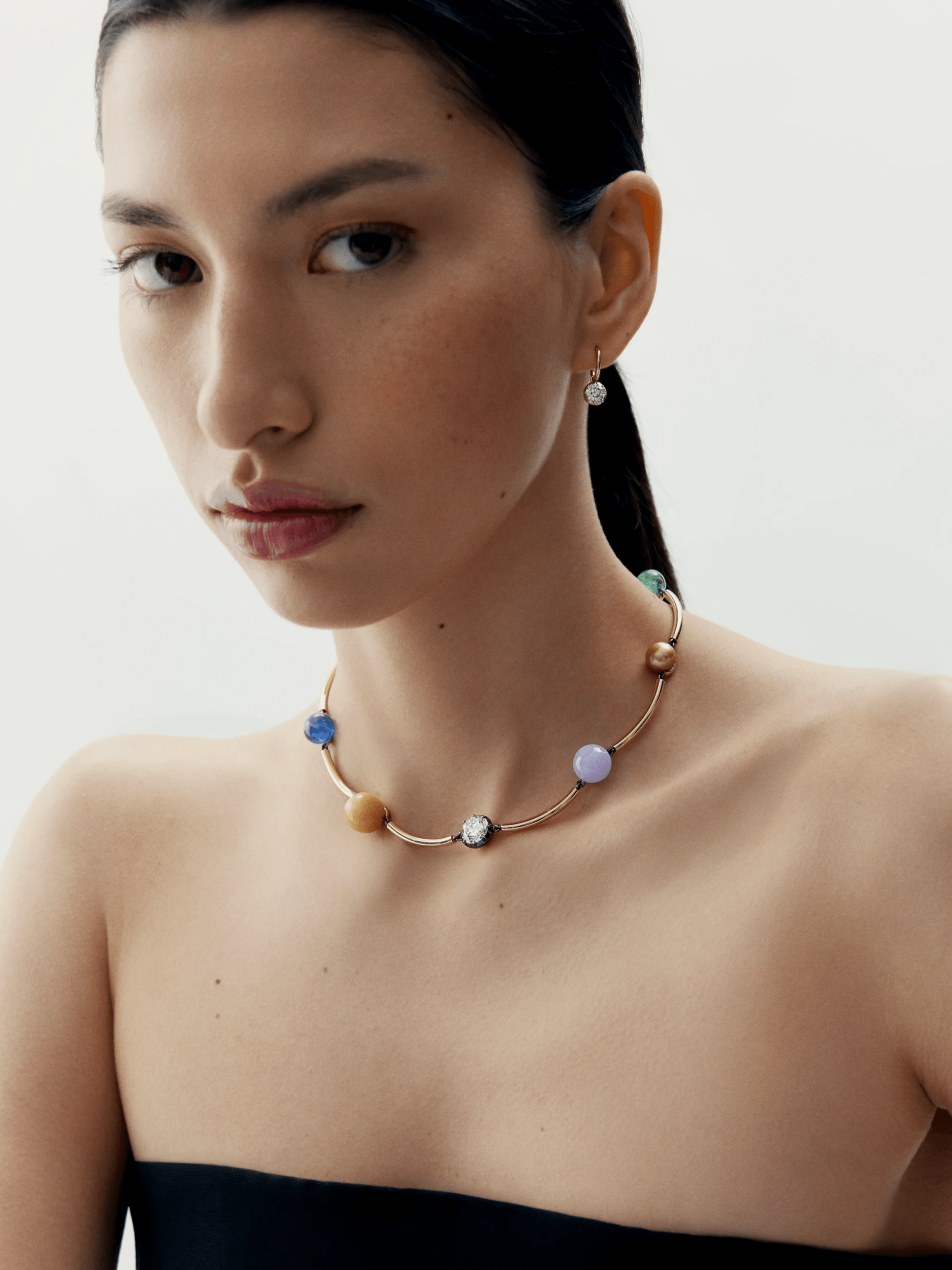 Close-up of a woman wearing a contemporary necklace featuring a curved gold band with colorful round gemstone accents, styled with a single diamond drop earring. View 3