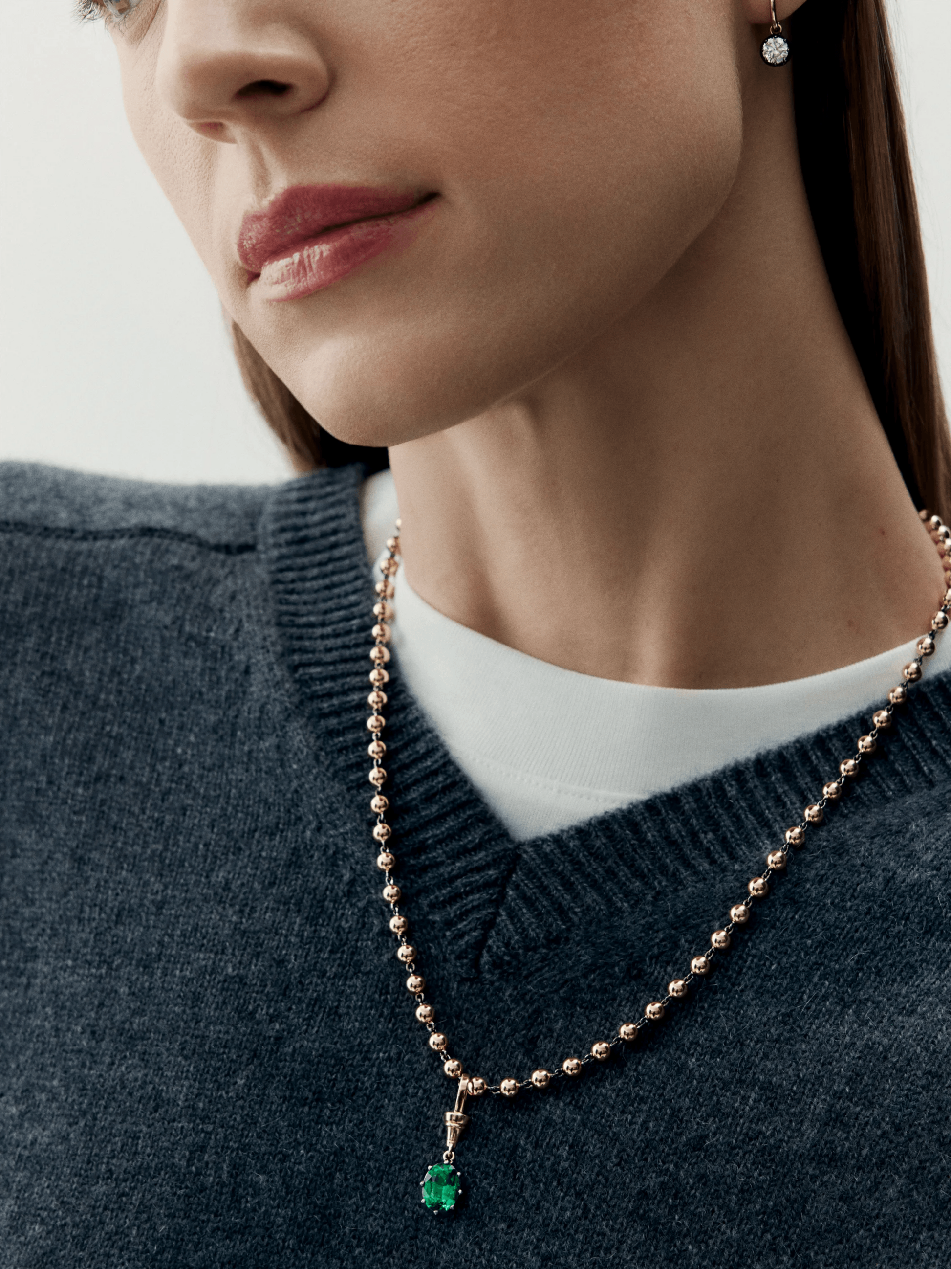 Close-up of a woman wearing a finely crafted rose gold ball chain necklace featuring a vibrant oval-cut emerald pendant, paired with a simple diamond stud earring. View 3