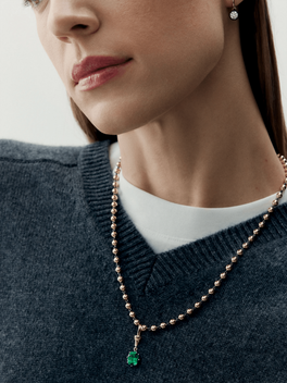 Close-up of a woman wearing a finely crafted rose gold ball chain necklace featuring a vibrant oval-cut emerald pendant, paired with a simple diamond stud earring.