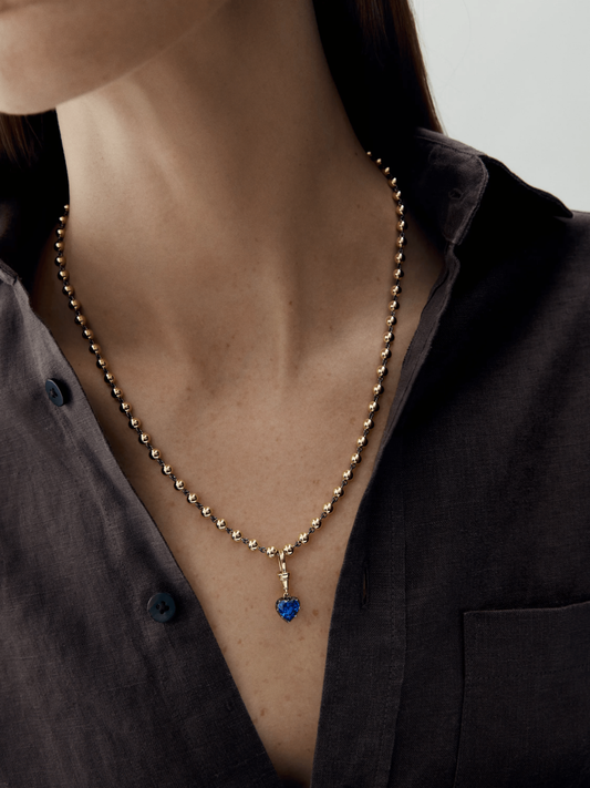 Close-up of a woman wearing a handcrafted gold ball necklace featuring a deep blue heart-shaped sapphire pendant, showcasing a blend of antique and contemporary design. View 3