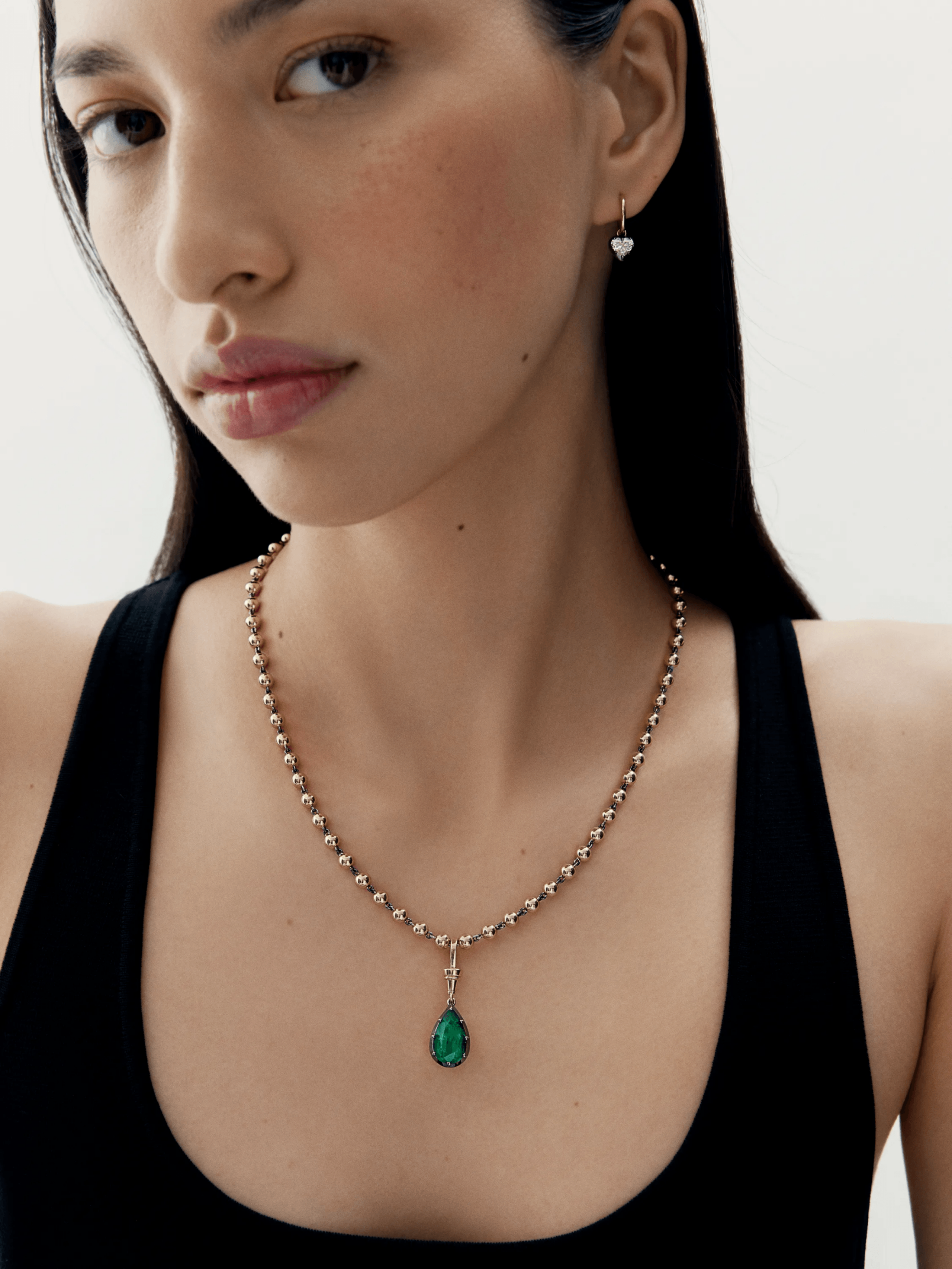 Close-up of a woman wearing a contemporary necklace featuring a pear-shaped emerald pendant set in a detailed gold chain, styled with minimalist makeup and a black top. View 3