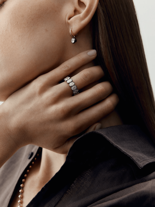 Close-up of a hand wearing an elegant oval diamond eternity band with millegrain detailing, styled with a minimalist diamond drop earring and dark clothing. View 3