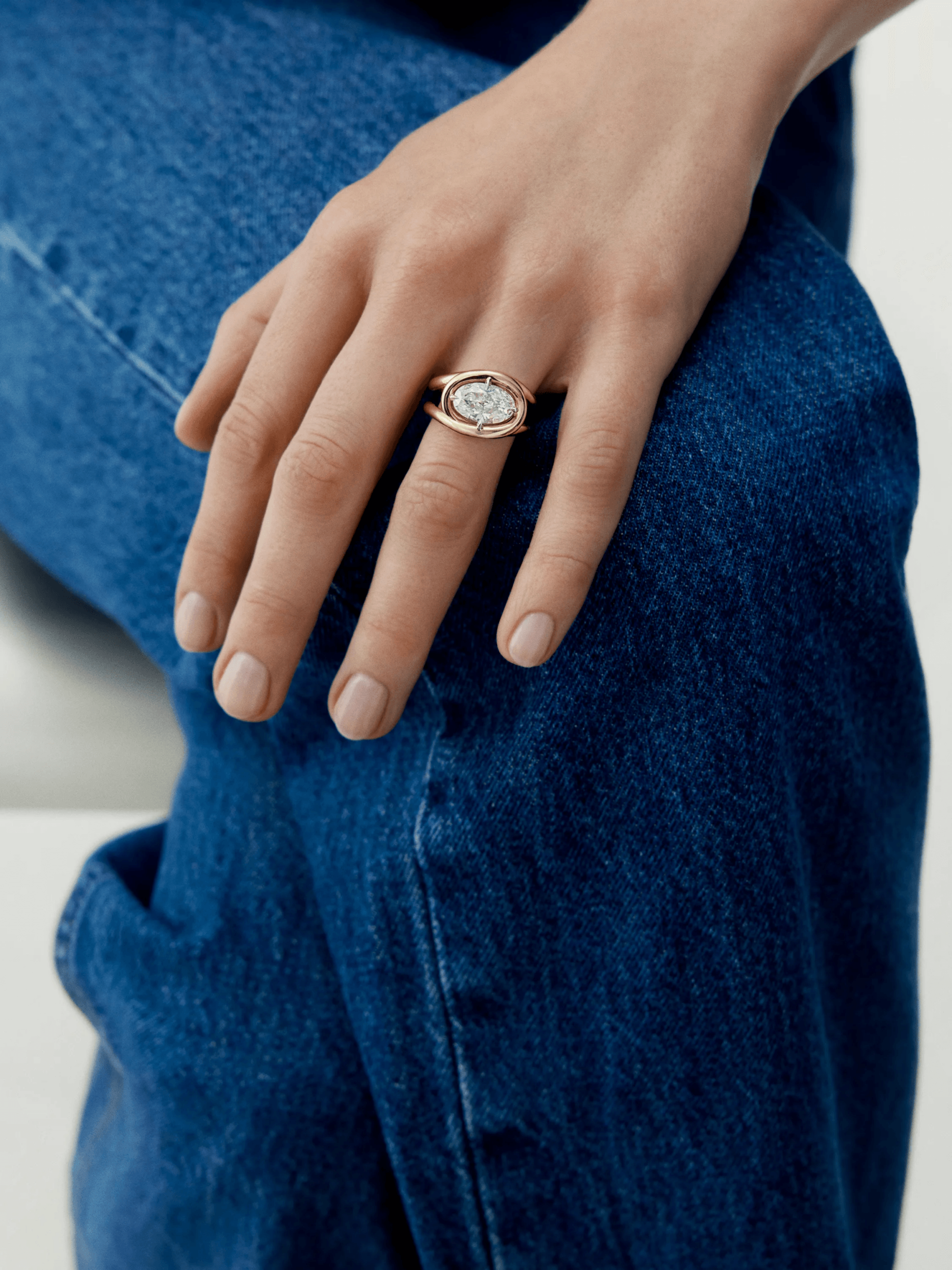 Hand wearing a rose gold ring with a 3.01ct oval diamond featuring a twisted band design, resting on blue denim jeans, showcasing contemporary craftsmanship. View 3