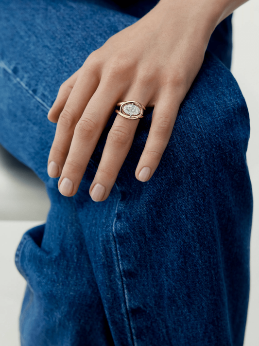 Hand wearing a rose gold ring with a 3.01ct oval diamond featuring a twisted band design, resting on blue denim jeans, showcasing contemporary craftsmanship. View 3