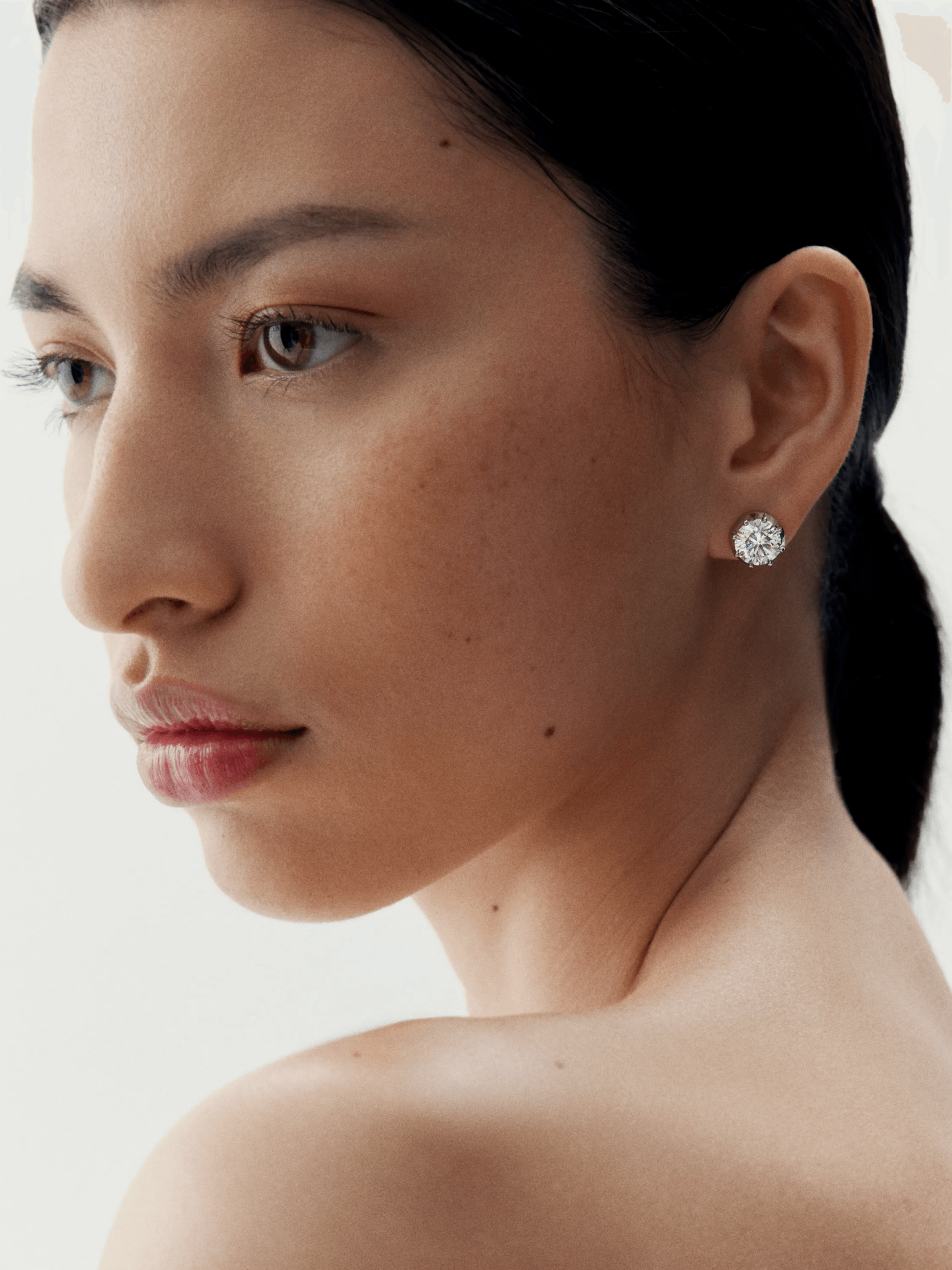 Close-up of a woman wearing elegant white gold stud earrings featuring a round diamond cut, showcasing refined craftsmanship with an antique-inspired contemporary design. View 3