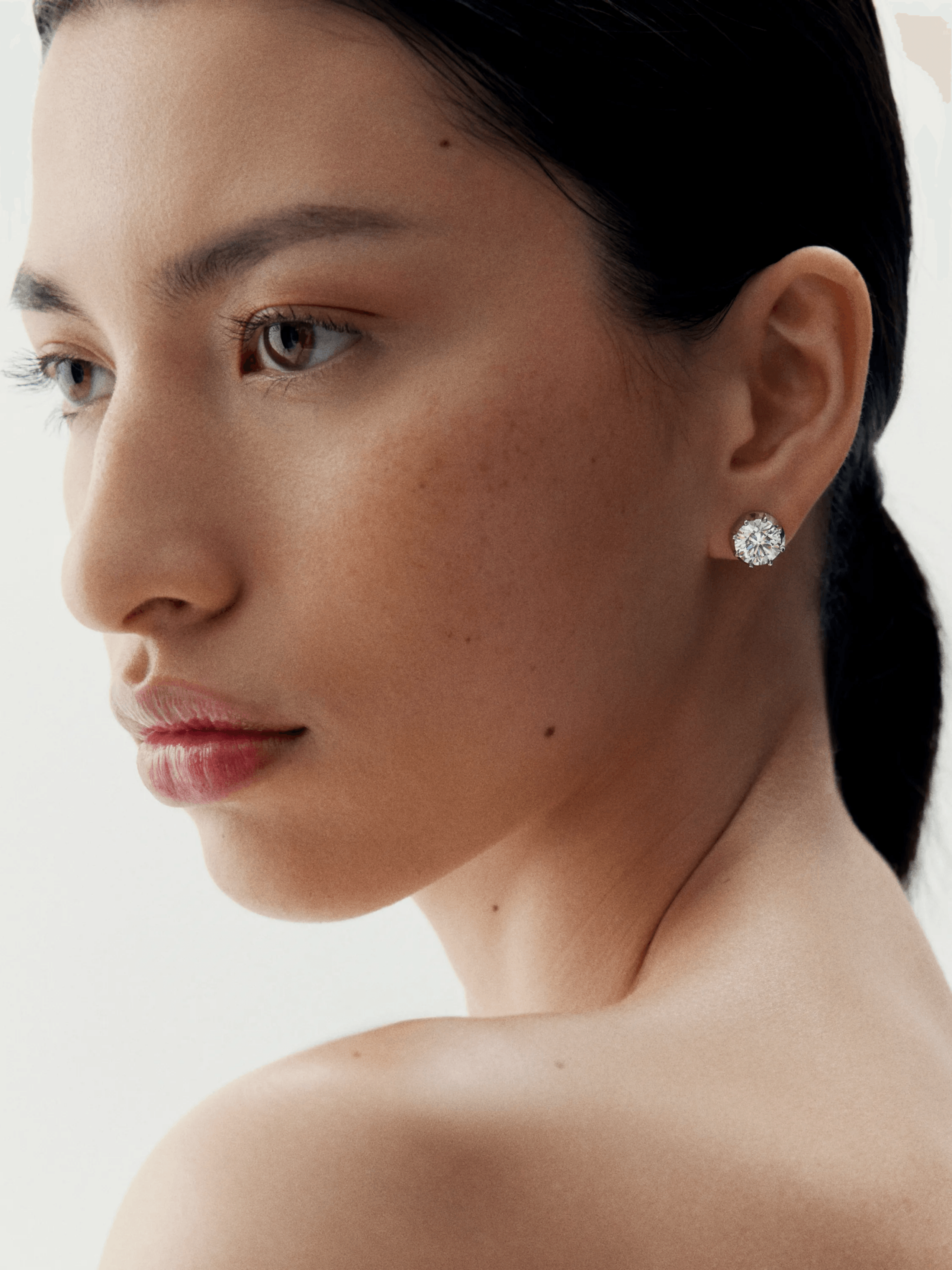 Close-up of a woman wearing elegant white gold stud earrings featuring a round diamond cut, showcasing refined craftsmanship with an antique-inspired contemporary design. View 3