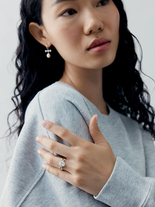 Woman wearing a cushion cut diamond ring with a distinctive bow-inspired setting, paired with a pearl drop earring and contemporary styling View 3