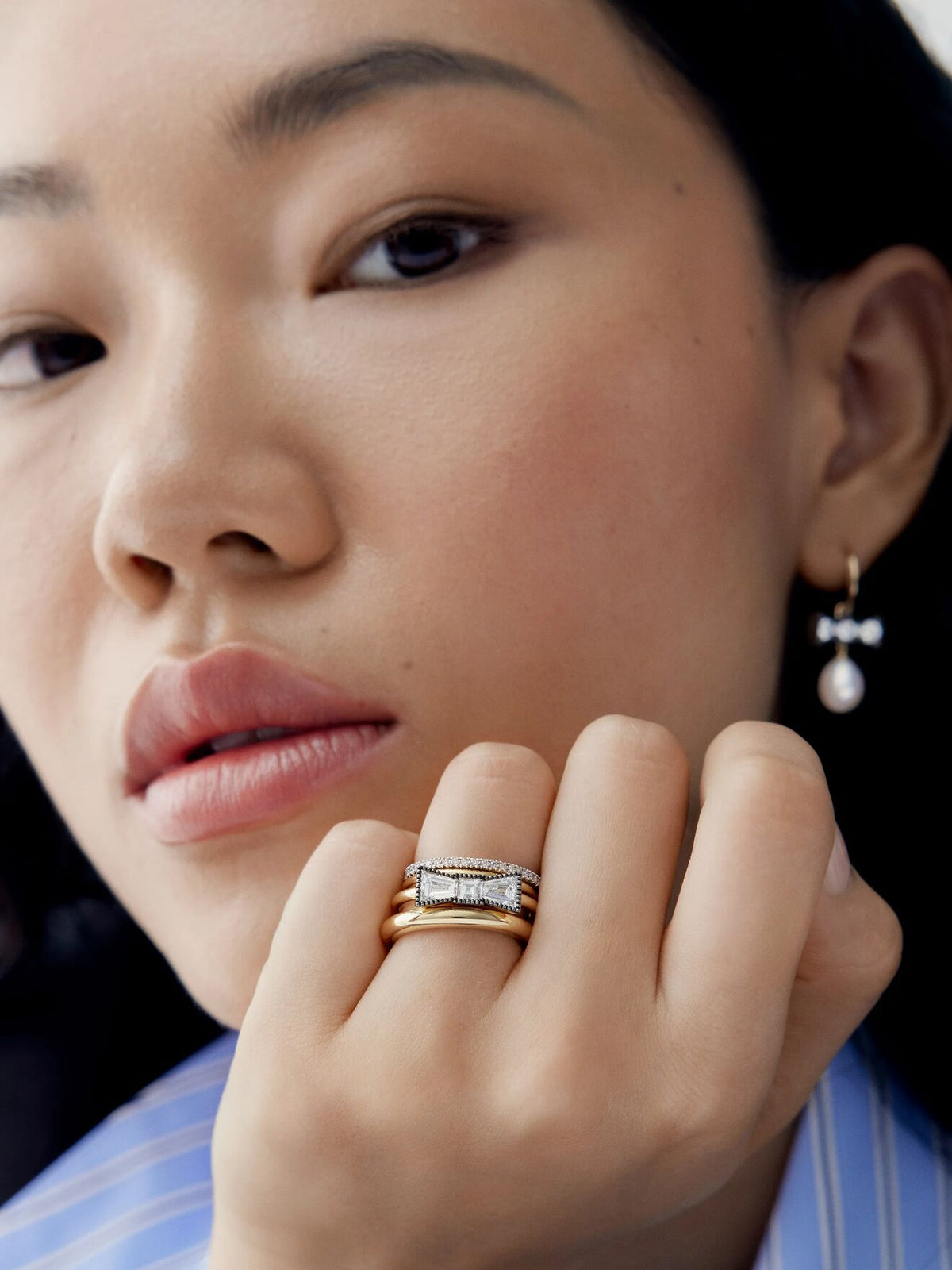 Close up portrait of a model wearing a diamond and blackened gold bow ring with layered gold bands and a pearl drop earring by Jessica McCormack View 3