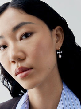 Close up portrait of a woman wearing a delicate diamond bow hoop earring with a suspended pearl, showcasing contemporary style and refined craftsmanship by Jessica McCormack