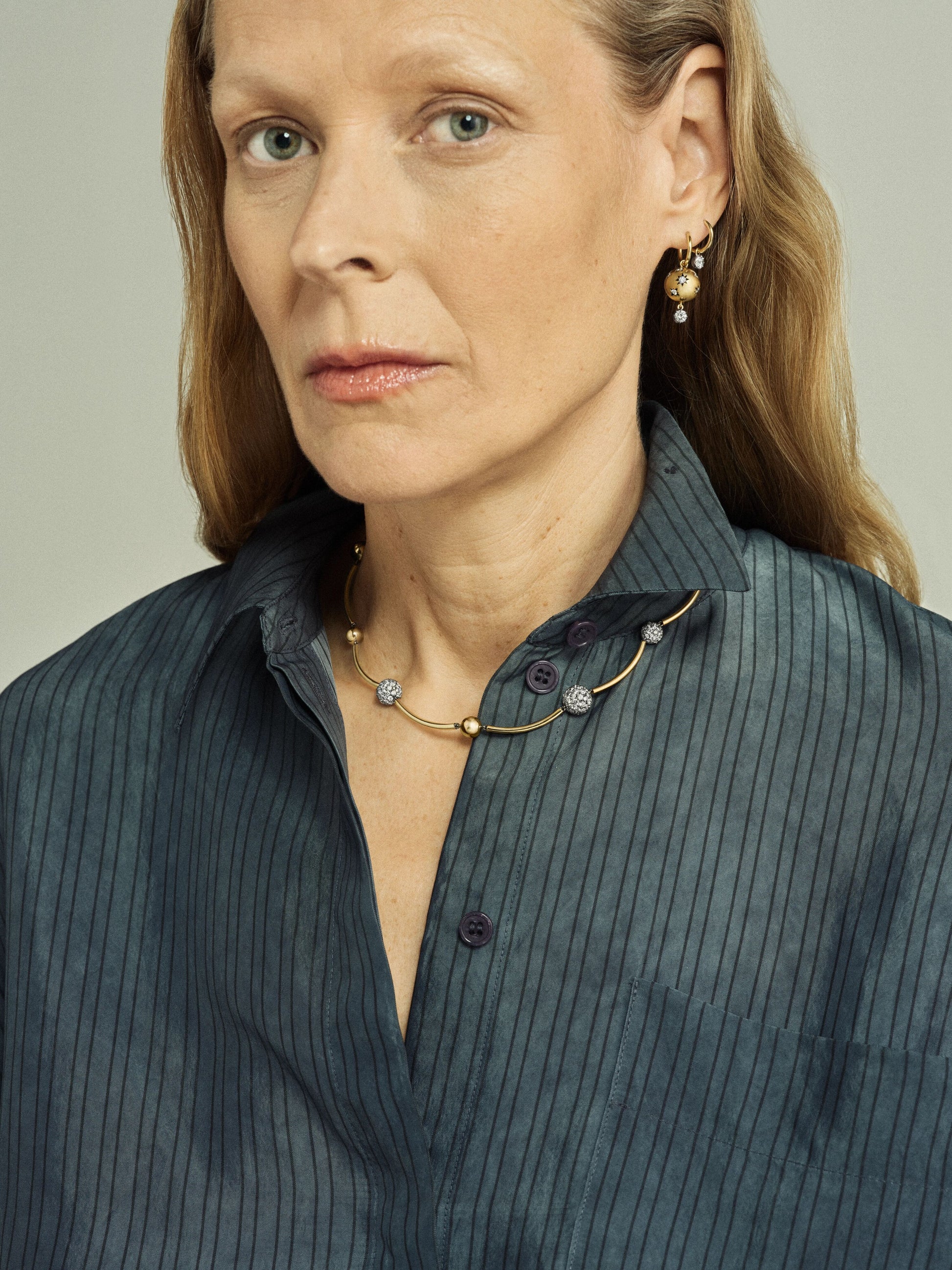 Close-up portrait of a woman wearing a yellow gold necklace with diamond pavé spheres, paired with matching delicate gold and diamond earrings, styled with a dark striped shirt. View 3